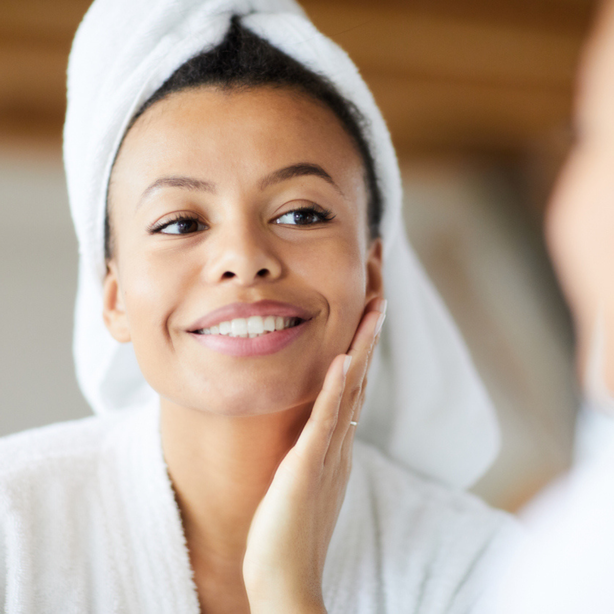 A black woman in a white robe smiling and looking at her skin.