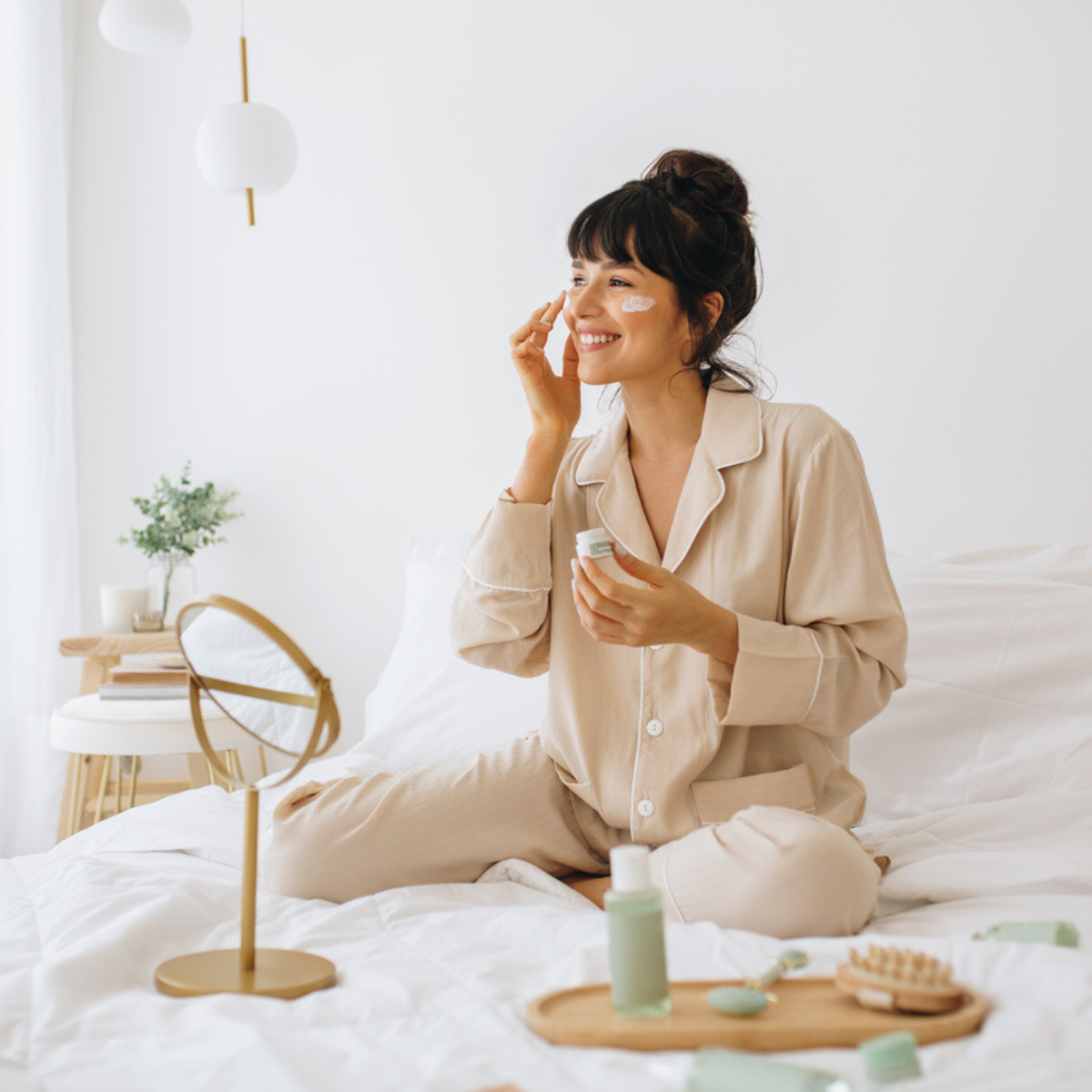 A woman in her pajamas sitting on her bed applying face cream.