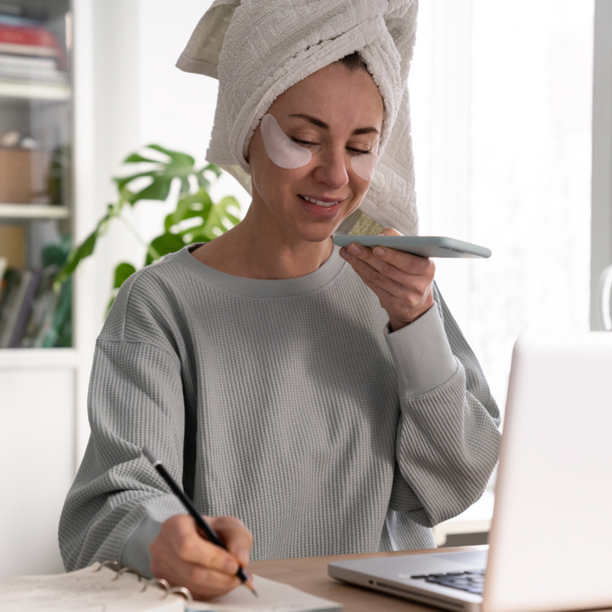 A busy woman doing skincare will taking a call and writing notes in front of her computer.