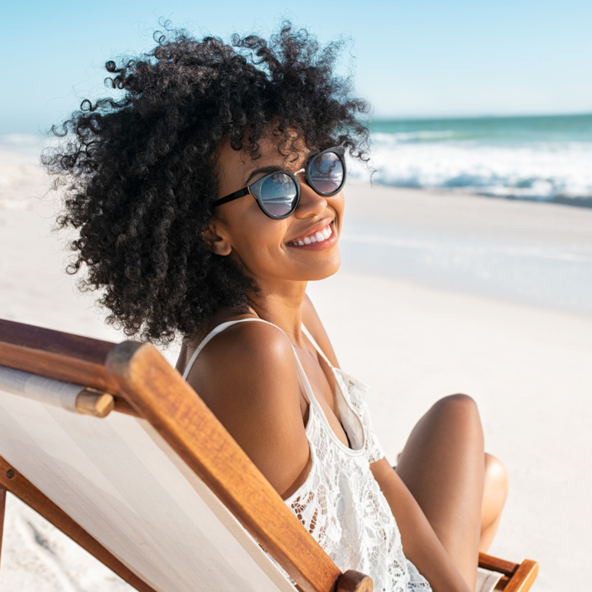 A black woman sitting on a lounge chair at the beach, smiling and enjoying the summer sun.