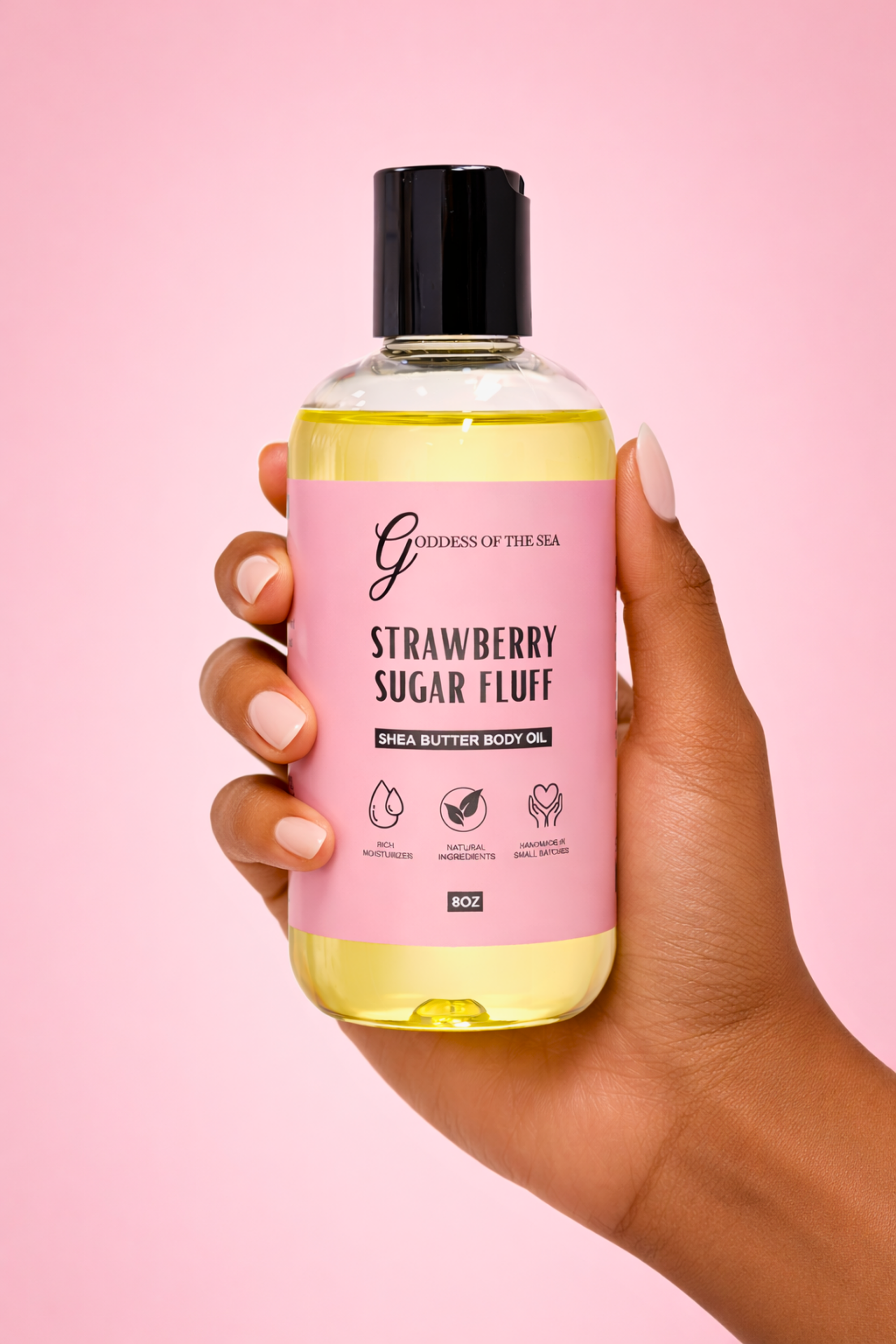 Hand holding a bottle of 'Strawberry Sugar Fluff' body oil with a pink label against a pink background.